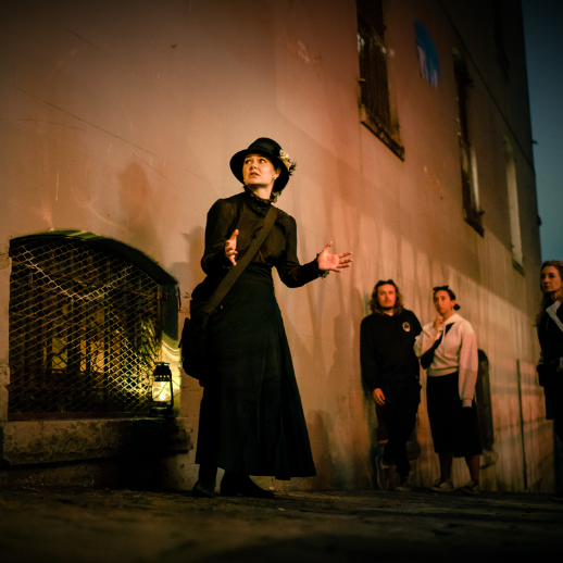 A sepia toned photo of the storyteller- a woman in 1800s garb, with tour guests gathered around as she tells a story