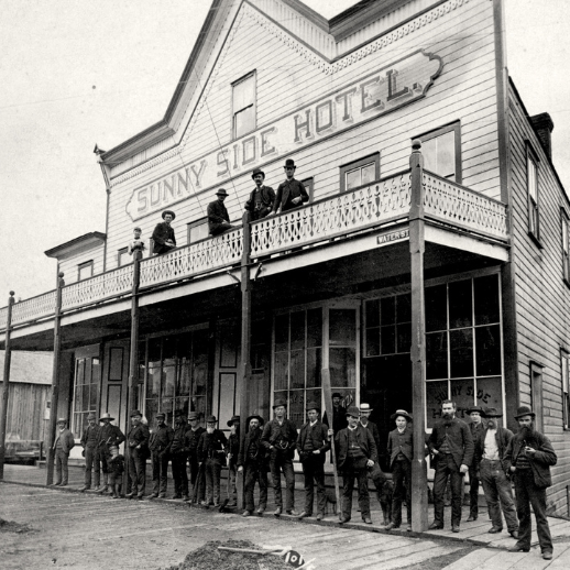 A black and white photo of the Sunny Side Hotel in Gastown with men dressed in old western wear posing for the photo on the ground floor and balcony.