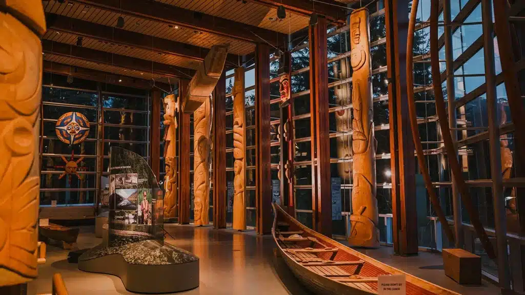 Top 10 Indigenous Tours & Experiences in Vancouver and BC 1 A photo of inside the Squamish Lil’wat Cultural Centre. There are floor-to-ceiling windows. It is night. There are totem poles and a canoe inside the centre.