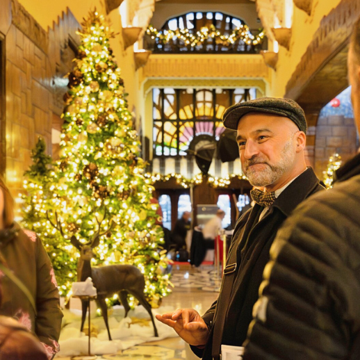 A photo of the lobby interior of the Marine Building during the holidays, with a lit up Christmas Tree. A guide smiles as he looks at his guests, who frame the photo.