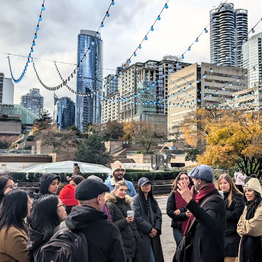 a guide gestures as a tour group smiles. Lights are strung up above with a view of buildings in the background