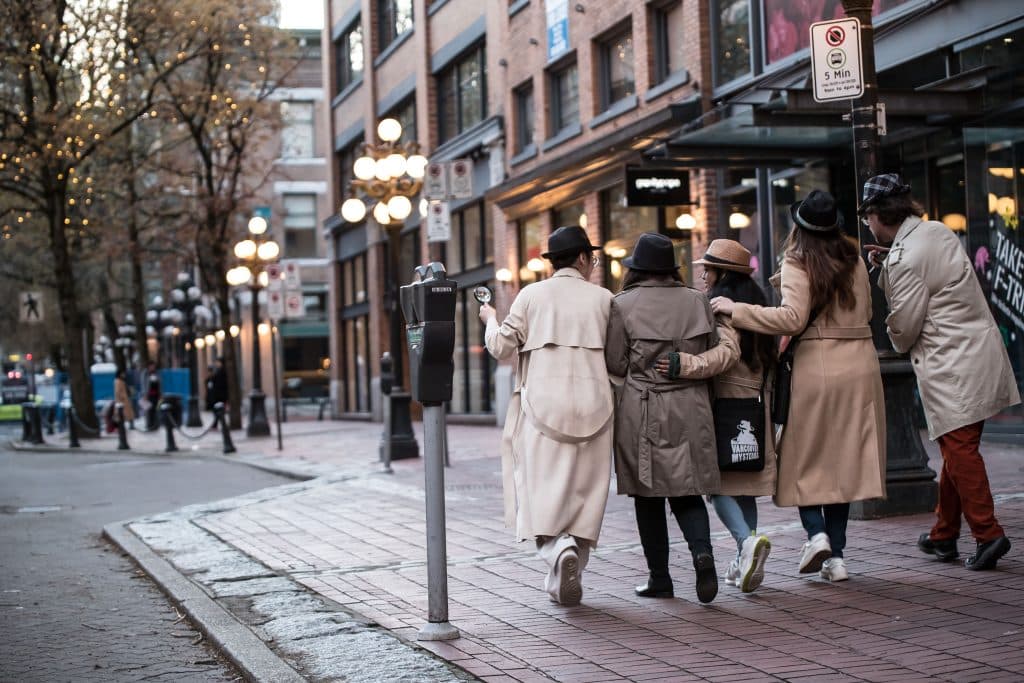 10 Actually Fun Things To Do In Vancouver for Young Adults 4 Four people wearing trench coats. Their backs are facing the camera. They walk down a cobblestone path in Gastown, Vancouver with their arms around each other. A streetlight is beside them.